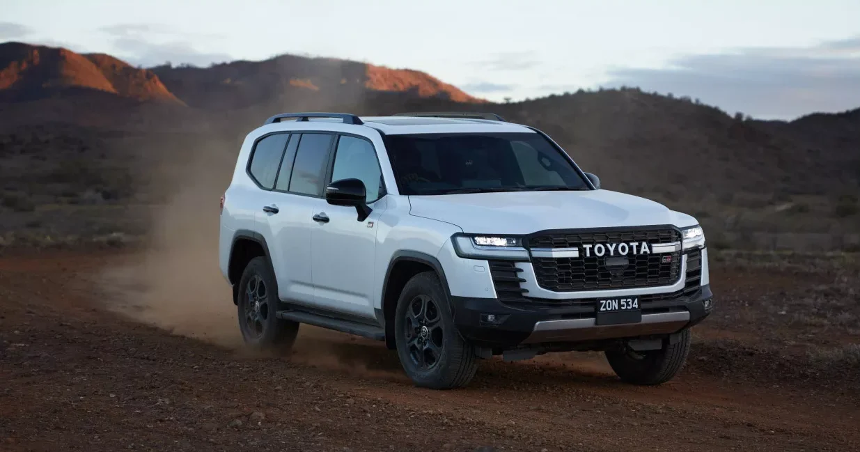 A white Toyota LandCruiser 300 Series driving on a dirt track through red hills