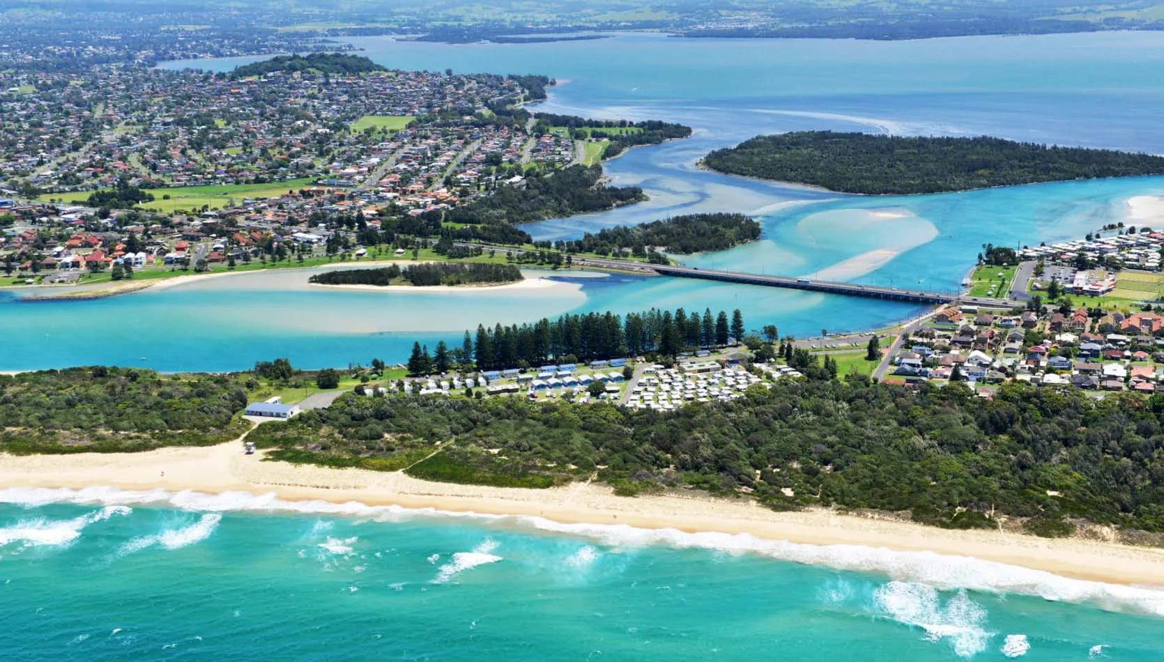 A sunny photo of Windang Beach Tourist Park, showing a grassy area with some trees and buildings in the background. A glimpse of the beach is visible in the distance.