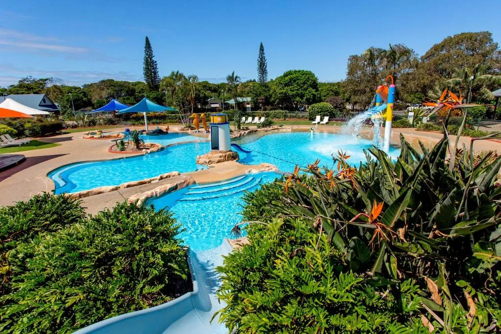 A photo of Coffs Harbour Holiday Park, showing a lush green lawn, a row of holiday cabins, and some trees. The sky is partly sunny.