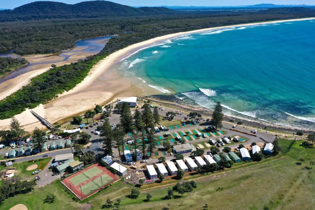 A scenic photo of Crescent Head Holiday Park, showing lush green grass, trees, and a glimpse of the ocean in the background. The park appears to be a camping or caravan park with possibly some accommodations visible.