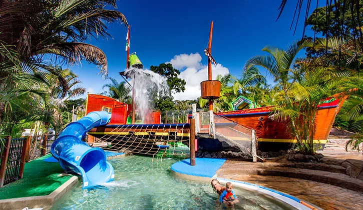 A photo of the NRMA Port Macquarie Holiday Park. It shows a lush green landscape with several holiday cabins visible, suggesting a relaxing and scenic vacation destination.