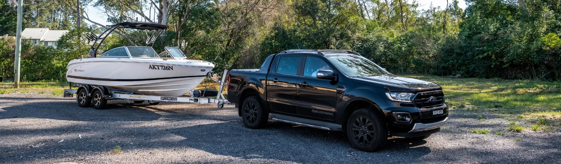 Black ute towing a white boat on a dual-axle trailer parked in a gravel area surrounded by trees