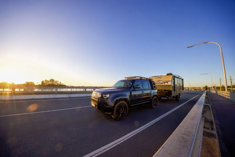 A Land Rover Defender towing an off-road camper across a bridge at sunset