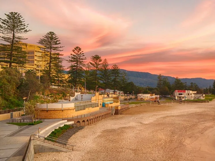 A photo of North Wollongong Beach in Australia. The image shows a sandy beach with calm ocean water and a partly cloudy sky.