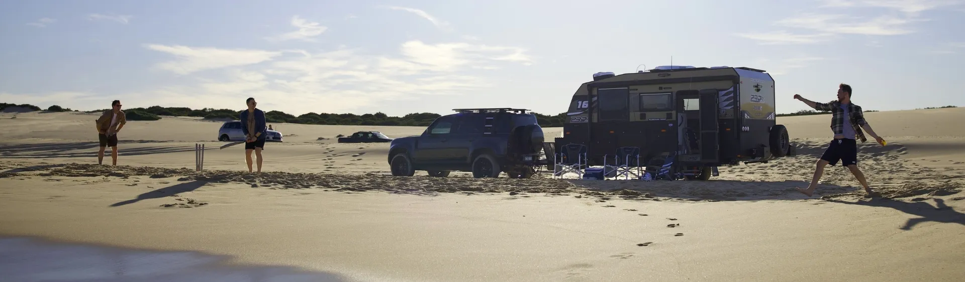 Beach scene with people playing cricket near a car and caravan.
