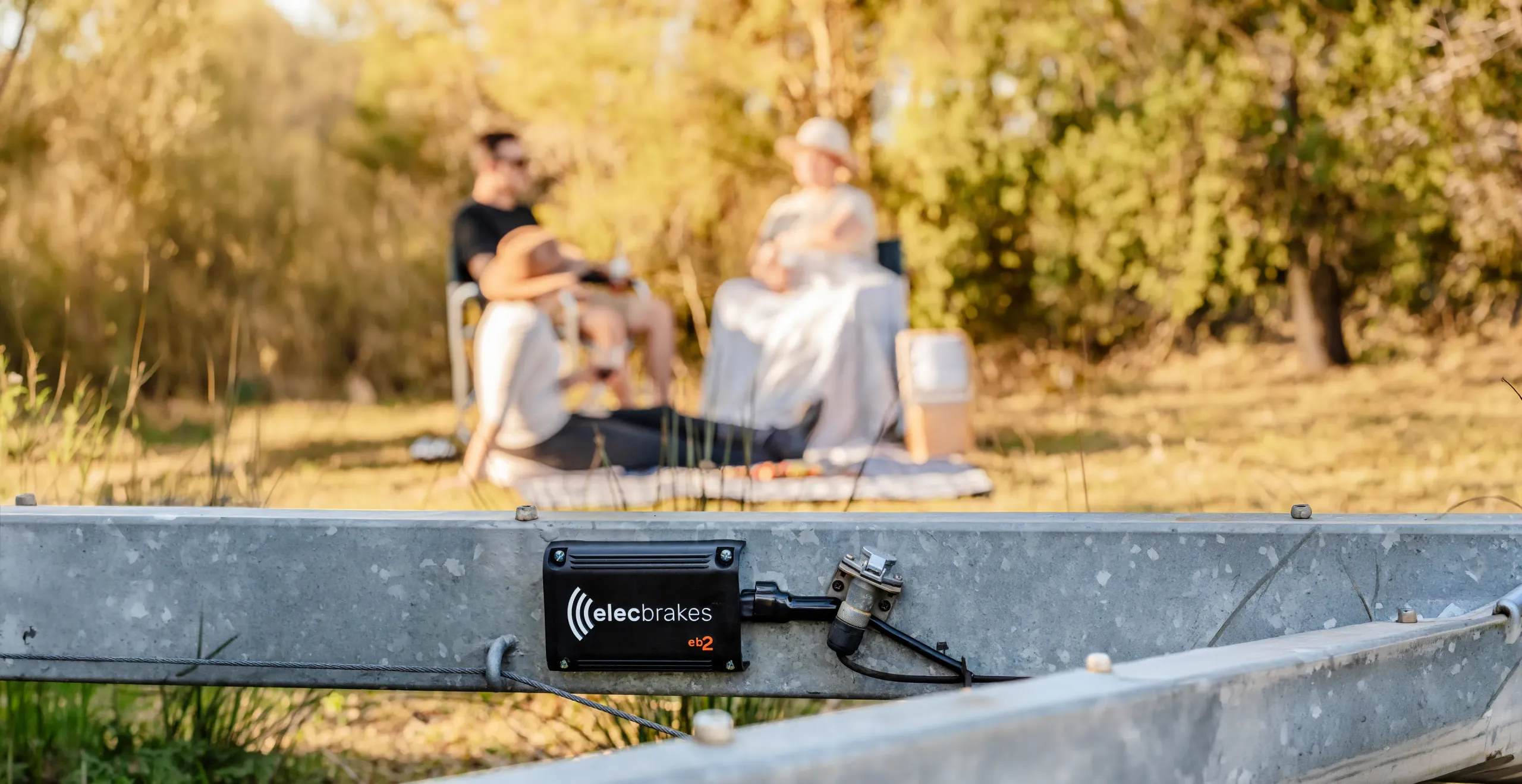 An EB2 brake controller mounted on a trailer drawbar, with a family relaxing on camp chairs in a sunlit bush clearing behind