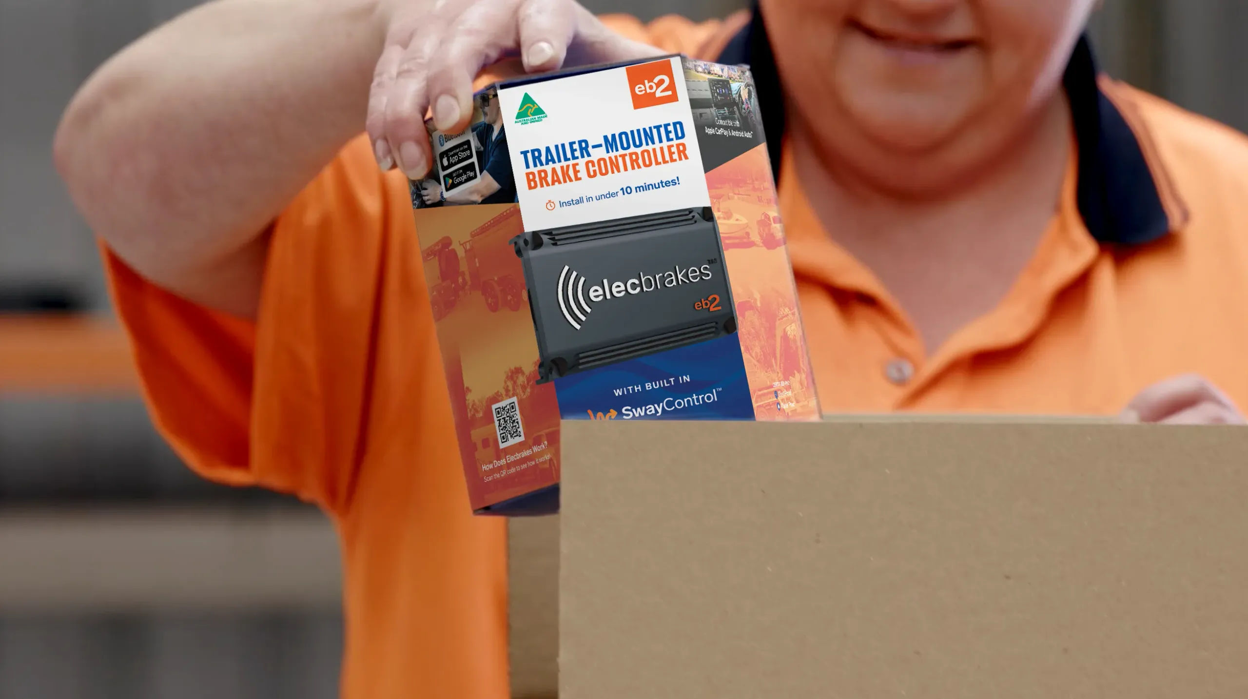 An Elecbrakes team member in an orange hi-vis polo lifting a boxed EB2 trailer-mounted brake controller out of a carton